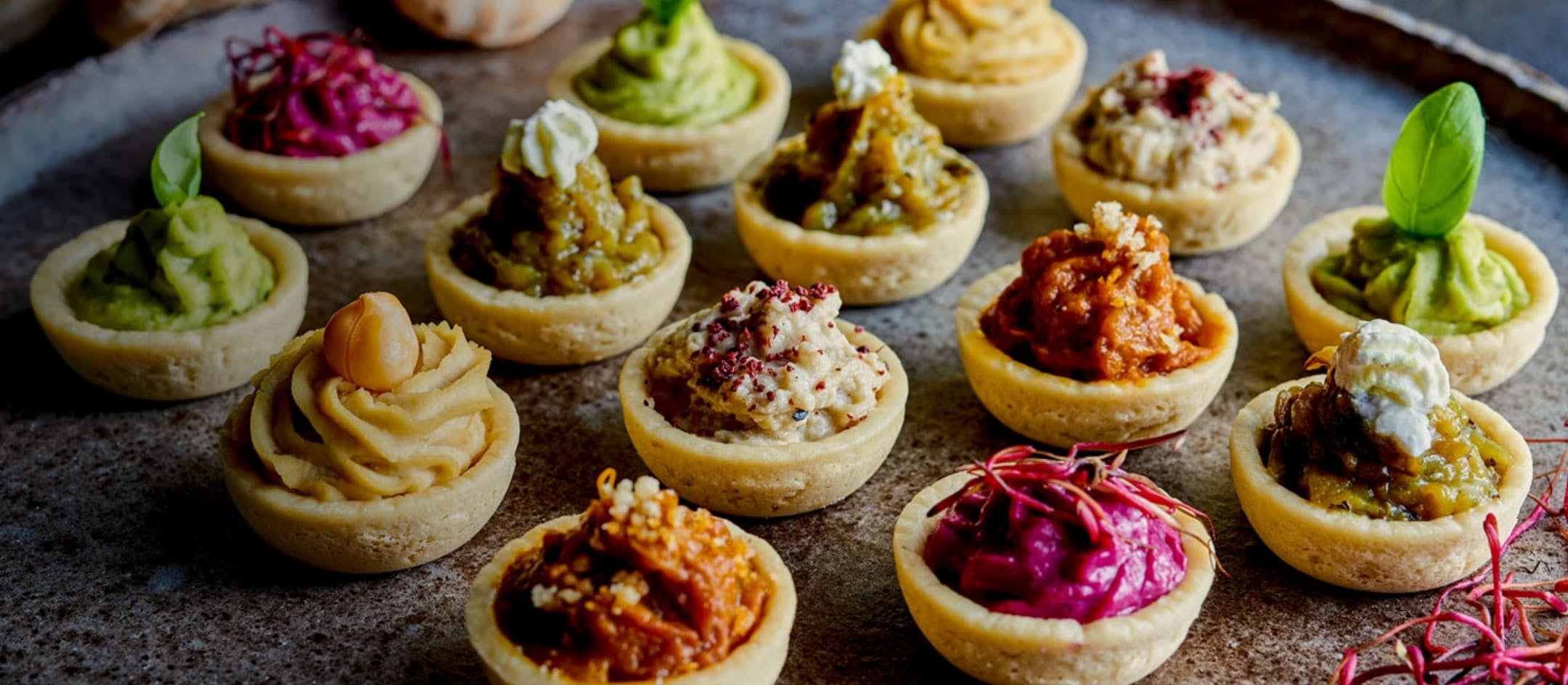 Close-up of various mini tartlets filled with different colorful Middle Eastern spreads, including chickpea, beetroot, and herb-based toppings, arranged on a rustic metal tray.
