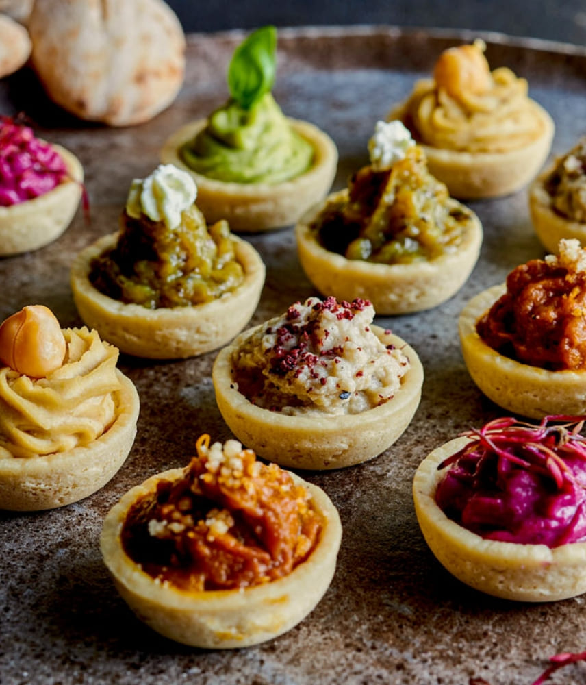 Close-up of various mini tartlets filled with different colorful Middle Eastern spreads, including chickpea, beetroot, and herb-based toppings, arranged on a rustic metal tray.
