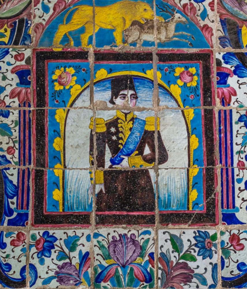 Detailed Persian tilework showing a historic royal figure in uniform, surrounded by floral patterns and a lion motif above, featuring vivid blue, yellow, and pink colors.