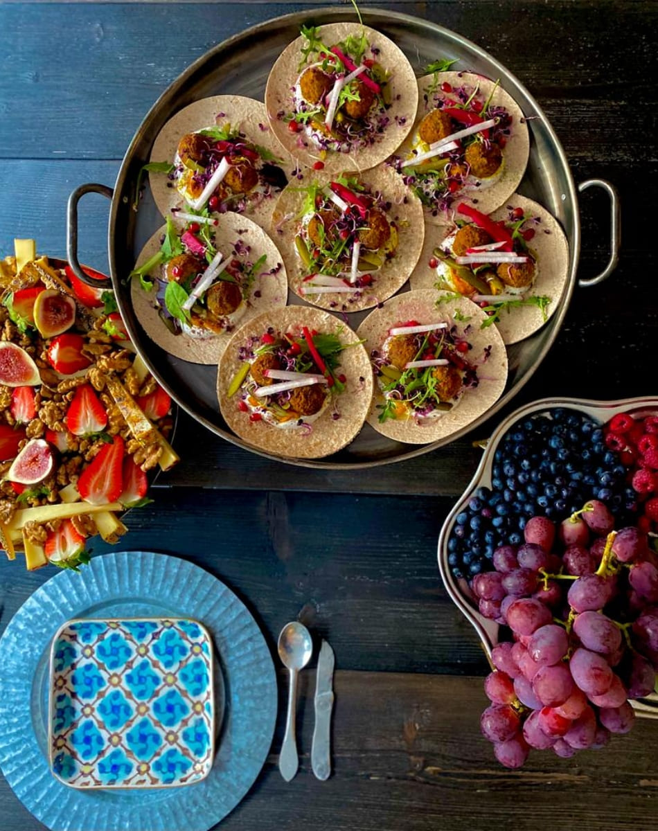 Overhead view of a vibrant table with falafel tacos on flatbread, a platter of assorted berries and grapes, and a tray with strawberries, figs, and cheese, all served on dark wood with a blue patterned plate.