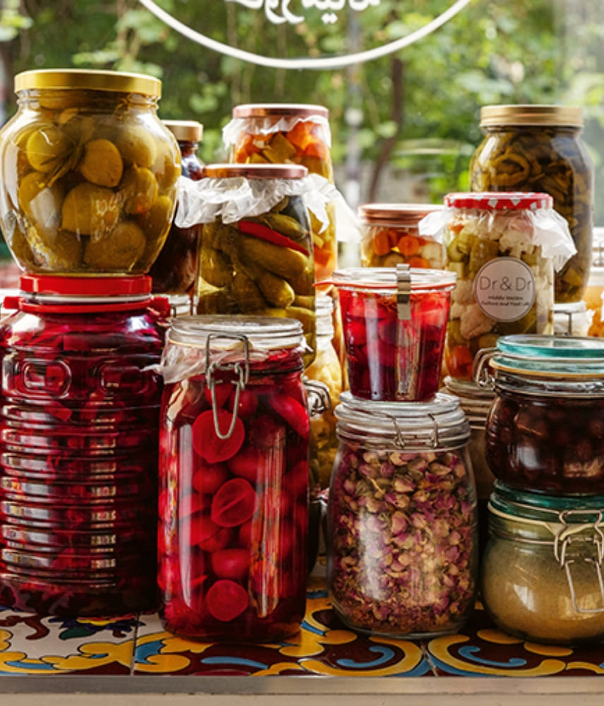 Various jars of pickled vegetables, pickles, and dried flowers on a decorative tile in front of a window, a perfect selection for delicatessen and pantry items.