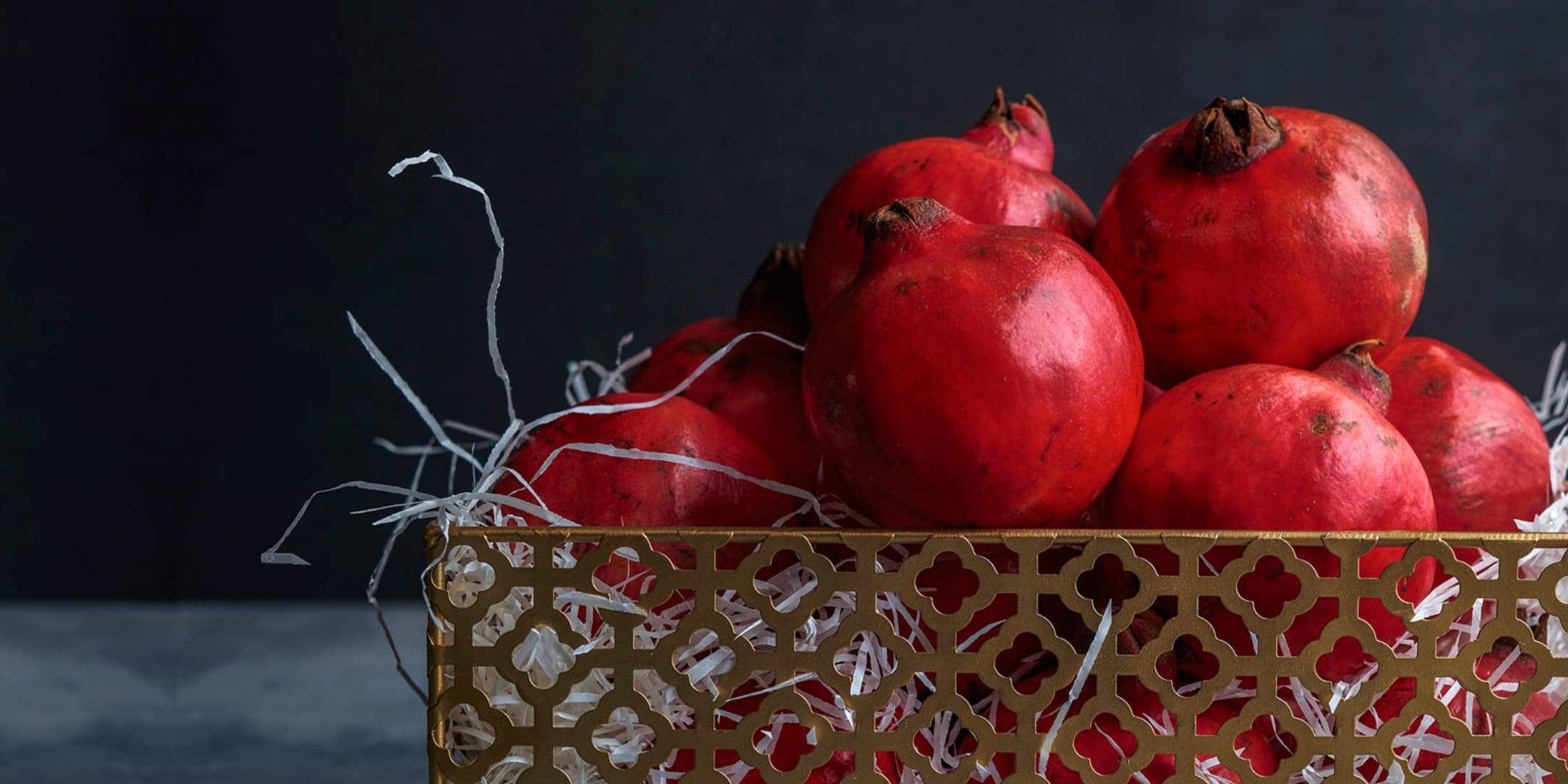 Close-up of a decorative golden basket filled with fresh red pomegranates and white filling material against a dark background