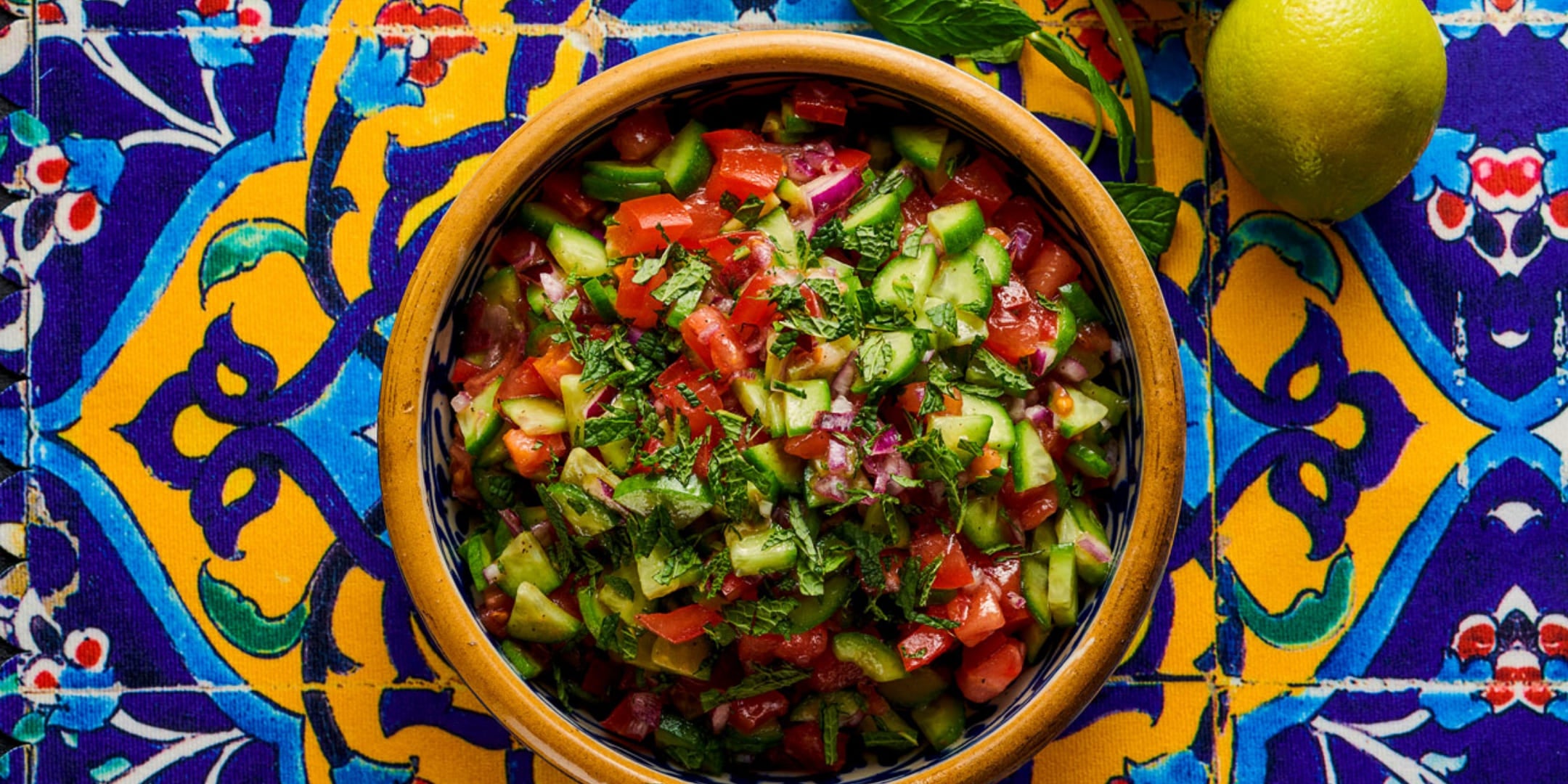 Close-up of a traditional clay bowl with Persian Shirazi salad of tomatoes, cucumbers, red onions and fresh herbs on oriental patterned fabric, decorated with a lime and mint.
