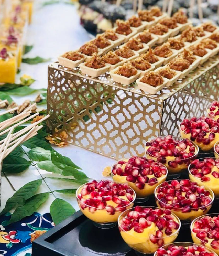 Close-up of Middle Eastern appetizers in small tart shells and dessert cups topped with pomegranate seeds, arranged on decorative trays with green leaves and wooden skewers.