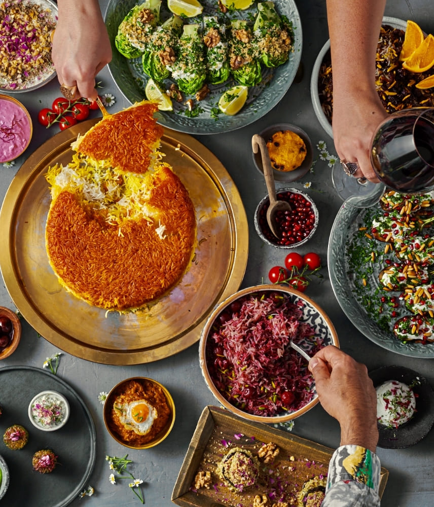Overhead view of a Middle Eastern feast with golden tahdig rice, colorful salads, dips, and hands serving food and holding a glass of red wine on a gray table.