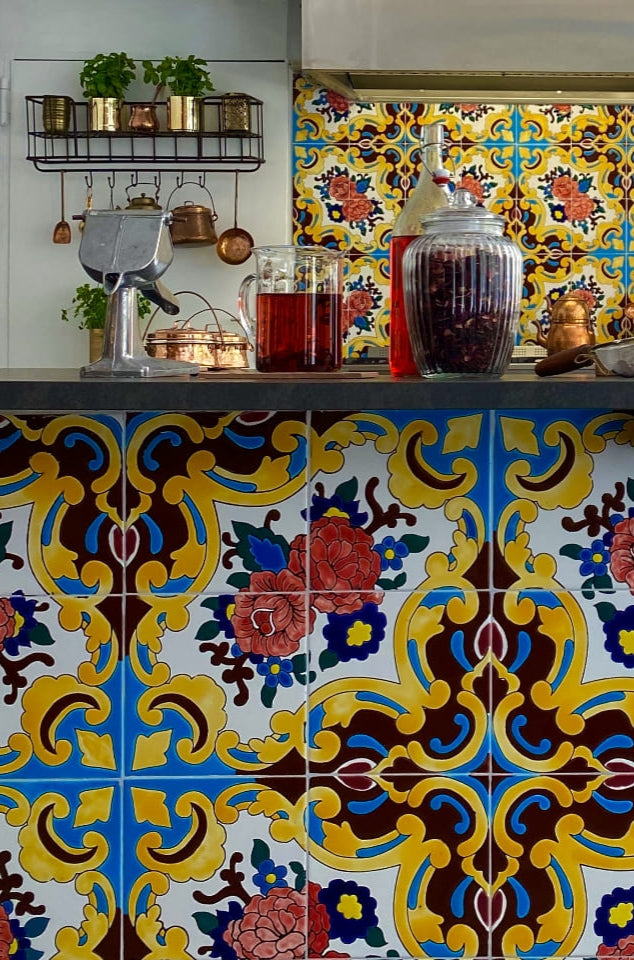 Kitchen counter decorated with vibrant floral and geometric patterned tiles, featuring jars, cooking utensils, and spices arranged neatly against a white cabinet backdrop.