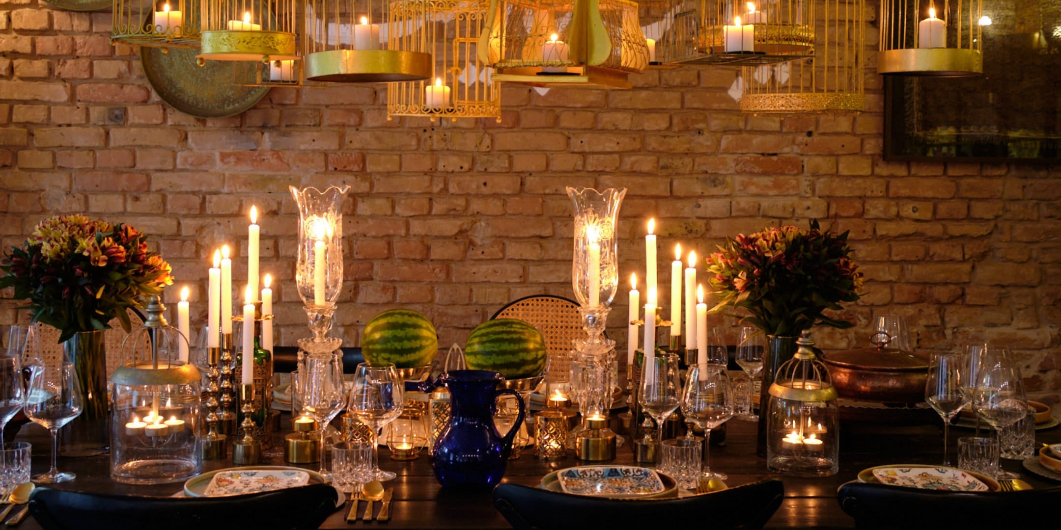 A beautifully set dining table with lit candles, glassware, blue ceramic pitcher, watermelon, and decorative birdcage chandeliers hanging above, against a brick wall backdrop.