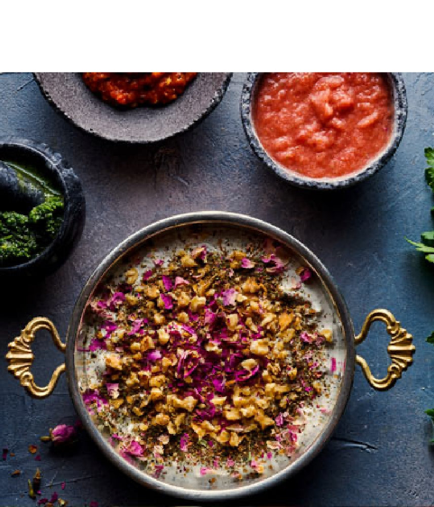 Bowl of creamy yogurt dip, garnished with chopped walnuts, dried herbs and rose petals, surrounded by small bowls of various sauces
