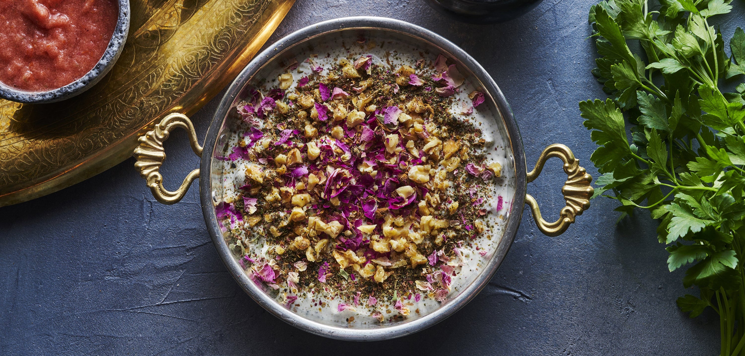 Bowl of creamy yogurt dip, garnished with chopped walnuts, dried herbs and rose petals, surrounded by small bowls of various sauces and fresh parsley