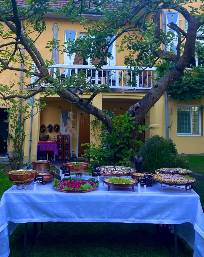 Outdoor buffet table covered with a white cloth displaying various dishes in copper and ceramic serving bowls, set in a garden with trees and a yellow two-story house in the background.