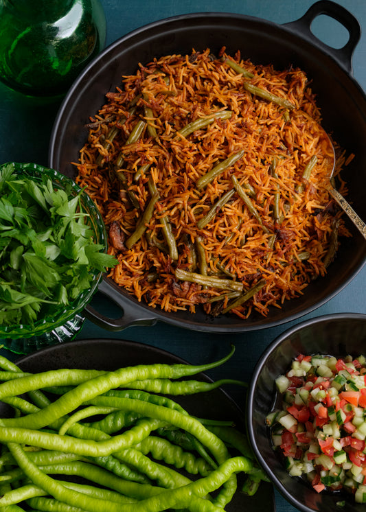 Black pan with Persian rice and green beans, surrounded by fresh parsley, green chilies and a salad of tomatoes and cucumbers