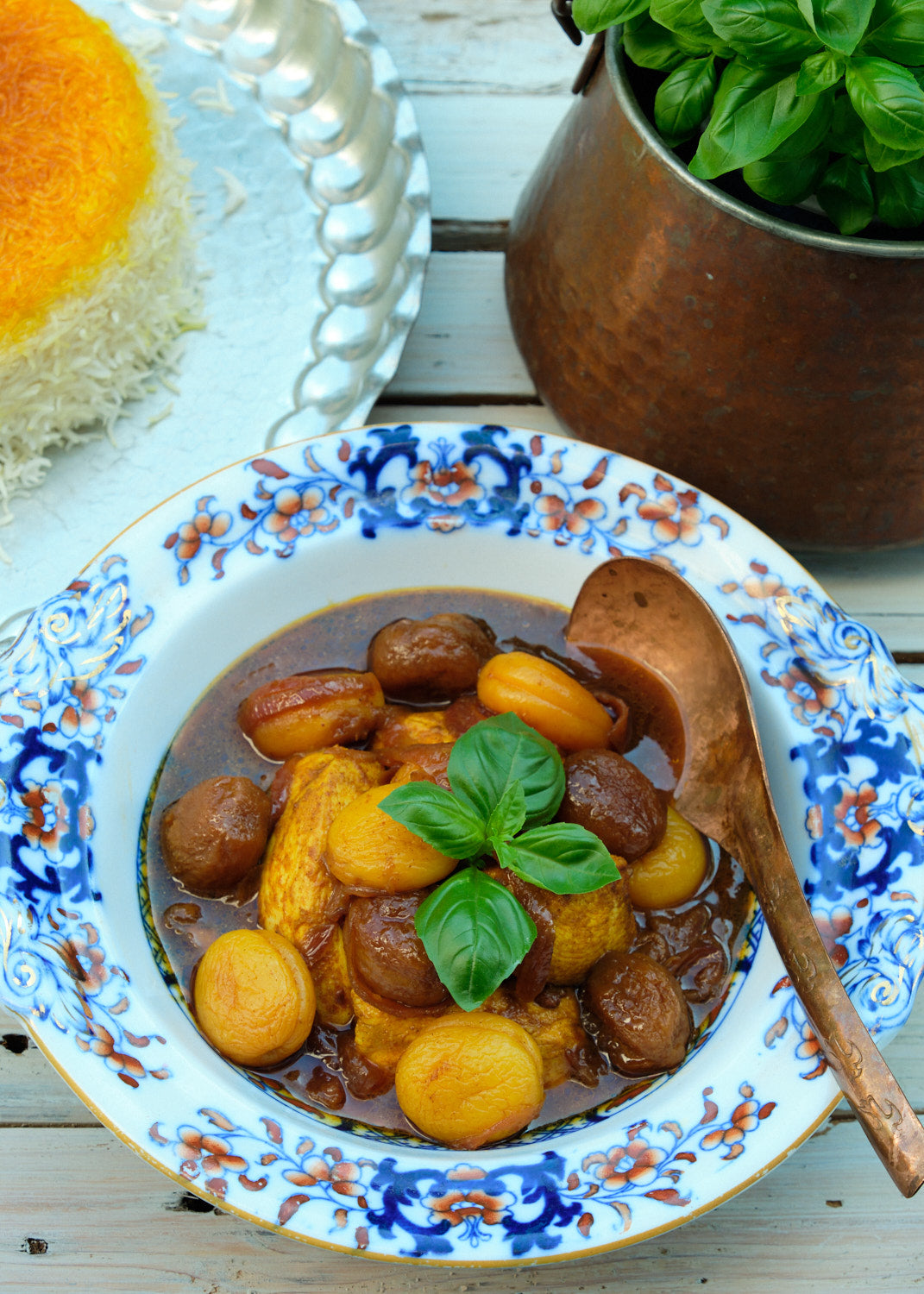 Braised chicken, dried apricots and plums, garnished with fresh basil, served on a decorated plate with saffron rice and a copper pot of basil