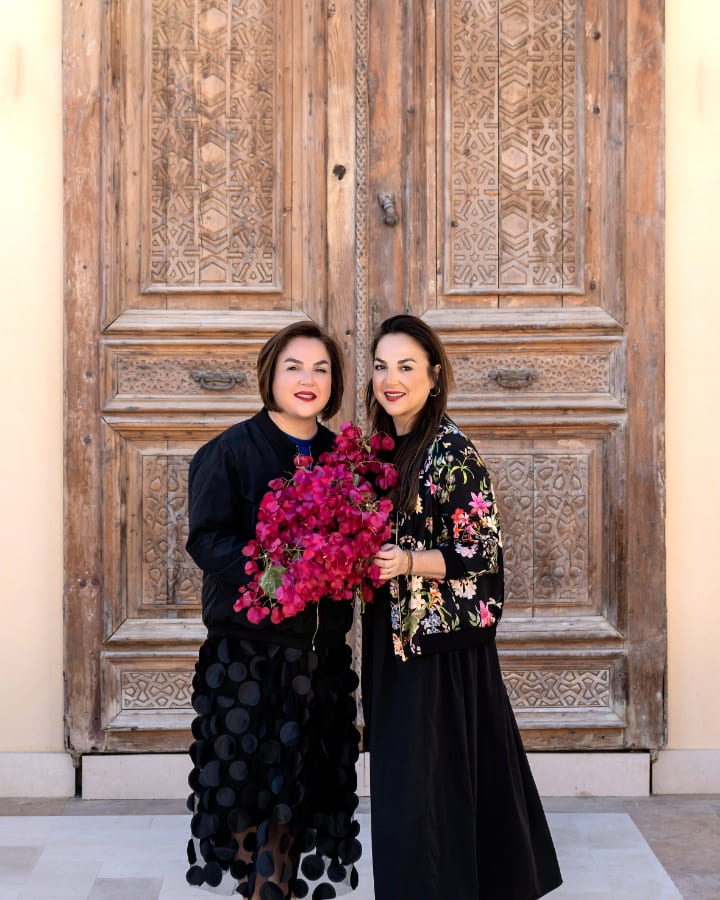 Two women standing in front of a large, intricately carved wooden door, smiling and holding a vibrant bouquet of pink bougainvillea flowers.