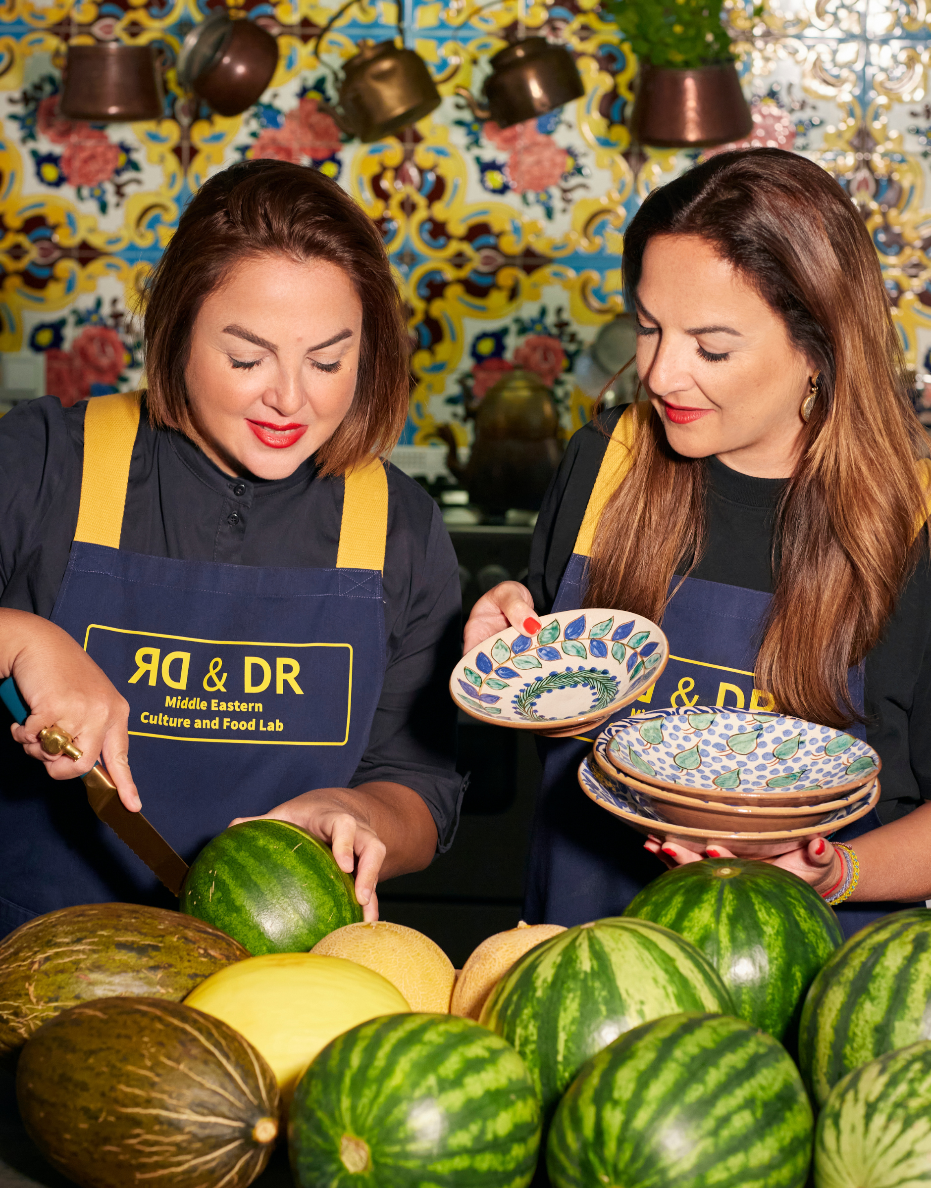 Two women in blue aprons cut a melon and hold hand-painted plates against a colorful, oriental-inspired background